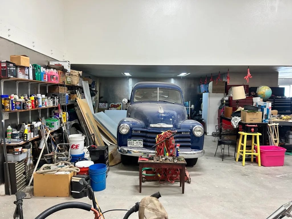 Vintage blue truck parked in a cluttered garage workshop with shelves of tools and supplies on both sides