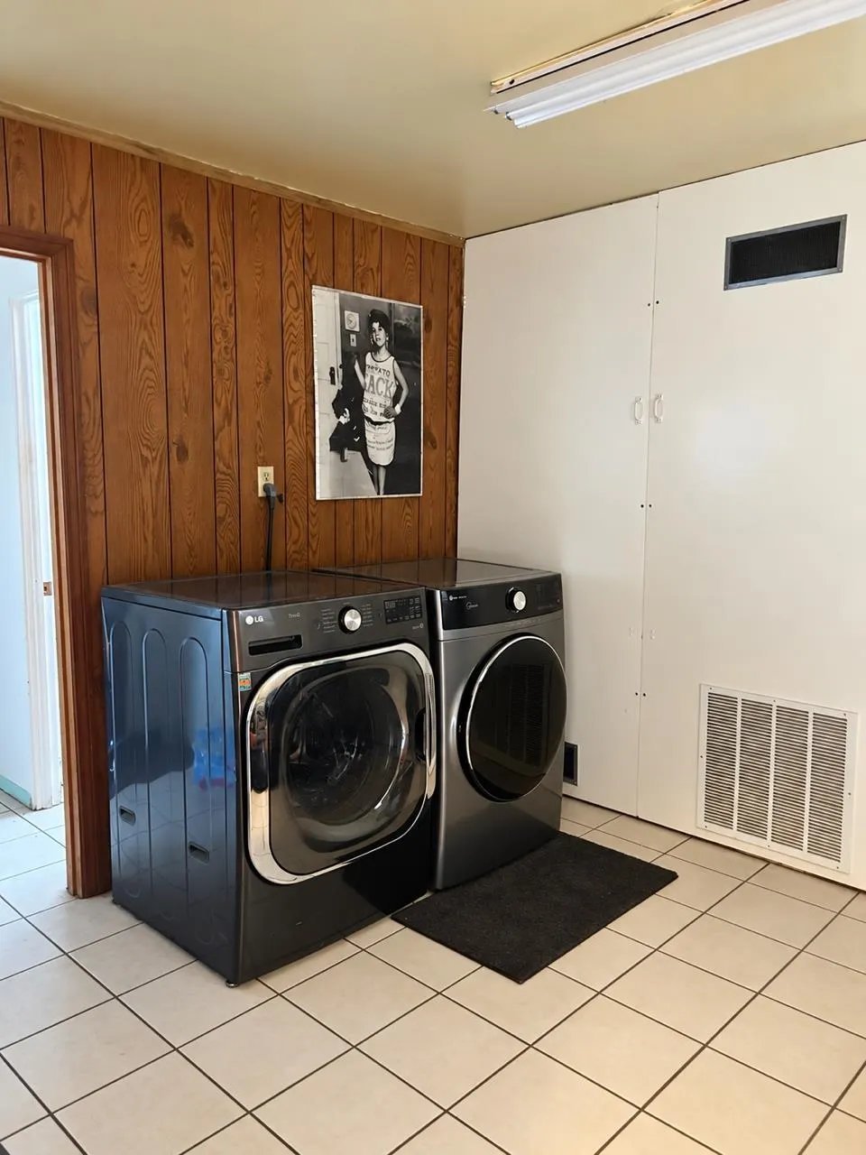 Laundry room with two black front-load washing machines on tiled floor, wood-paneled wall, and white cabinetry