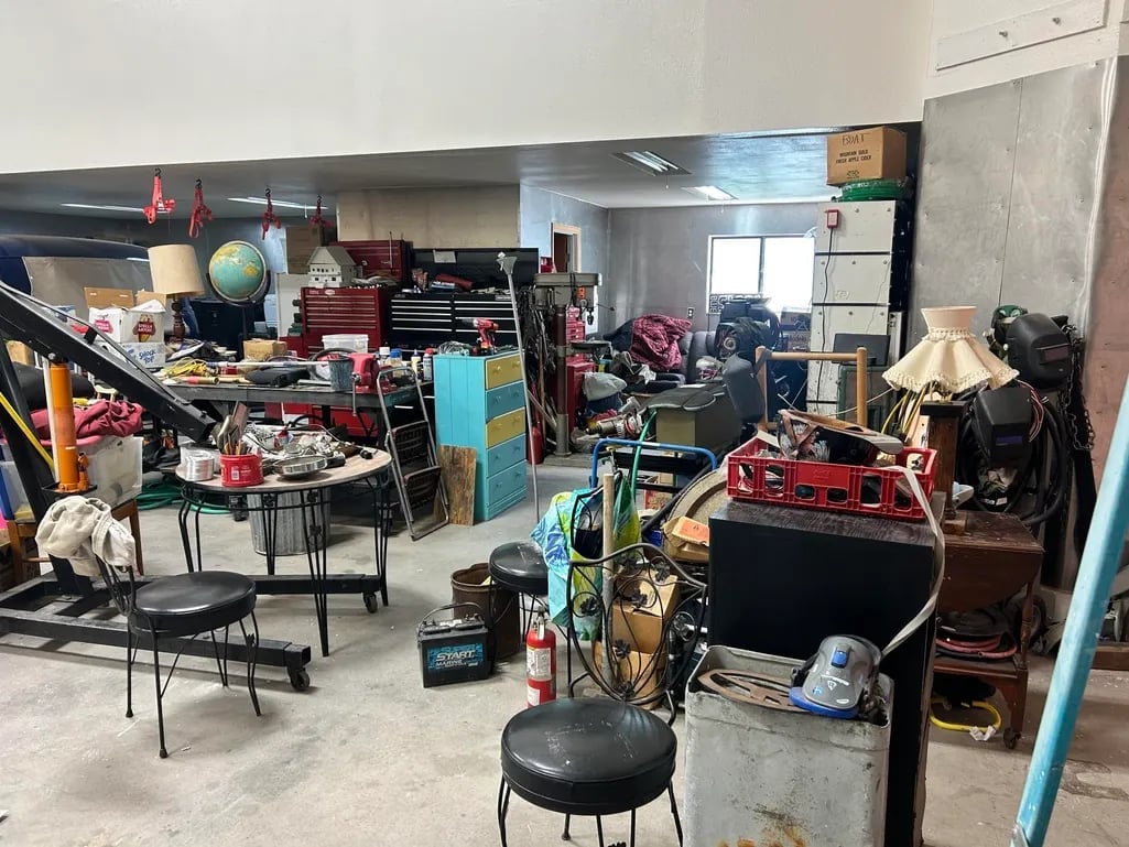 A cluttered garage filled with miscellaneous items, tools, furniture, and storage boxes scattered throughout the concrete floor space.