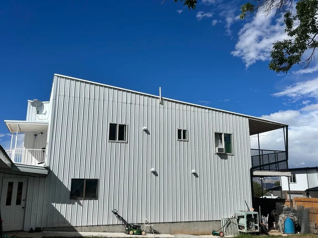 Modern white corrugated metal building with multiple windows and an open carport, set against a blue sky with clouds