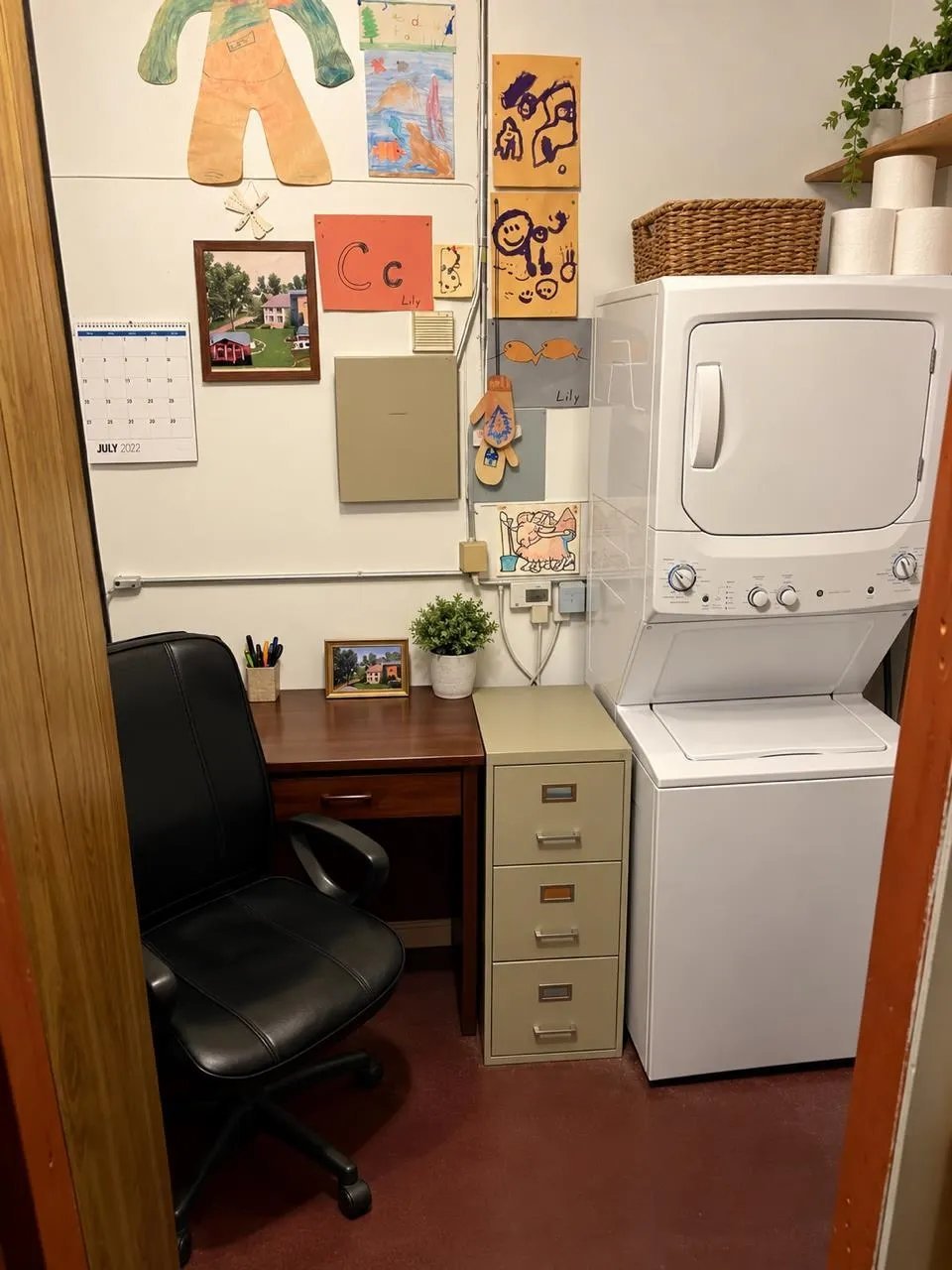A small laundry room with a stackable washer-dryer unit, wooden desk with black chair, filing cabinet, and childrens artwork displayed on the wall