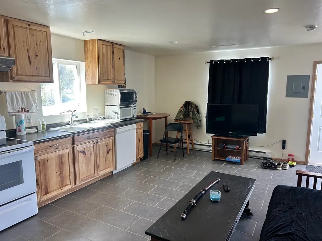 Open-concept kitchen and living room with wooden cabinets, tile flooring, black window treatment, and furniture arranged in the space.