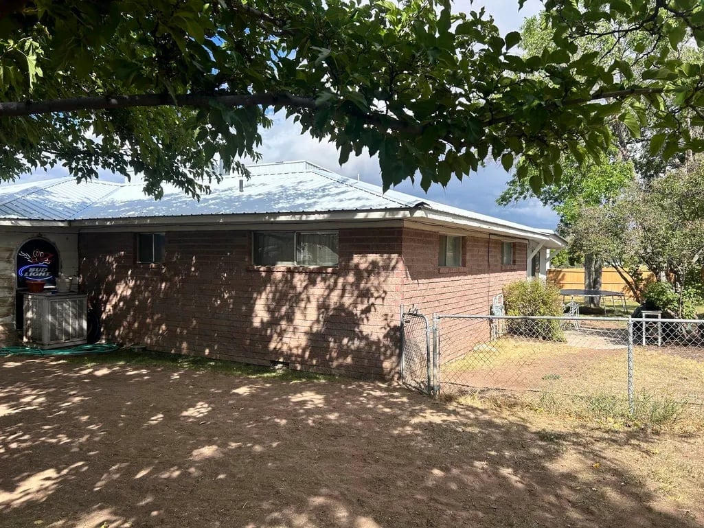 Brick residential house with metal roof surrounded by green trees and a metal fence on dry ground