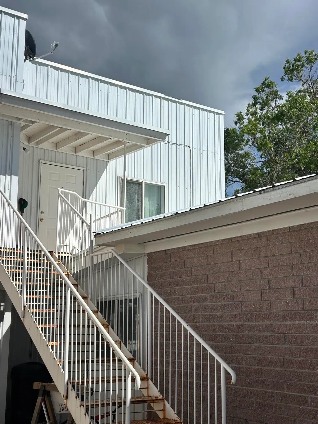 Modern white metal-sided structure with rooftop deck, accessed by metal staircase with railings against brick wall.