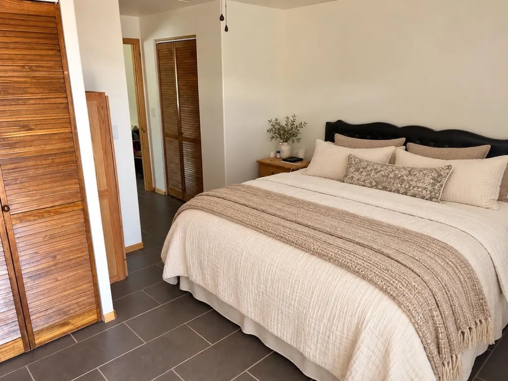 Modern bedroom with queen bed, cream bedding, black headboard, wooden doors, potted plant, pendant light, and dark tile flooring