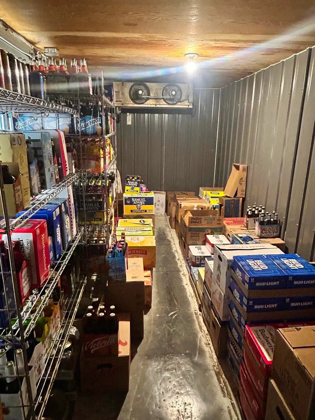 Interior of a storage unit or warehouse with metal shelving on left side stocked with merchandise, cardboard boxes stacked on floor, and cooling units mounted on ceiling