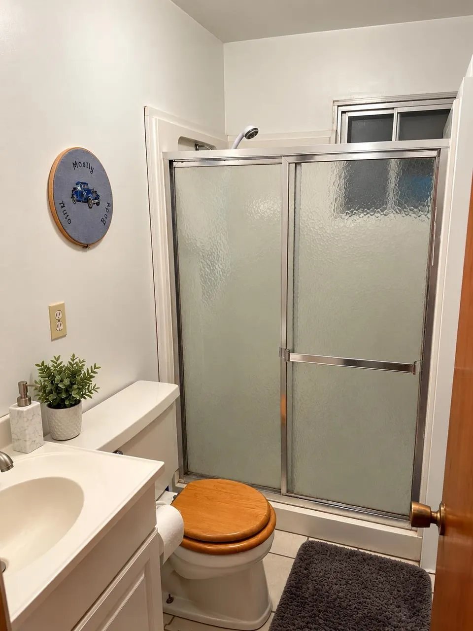 Modern bathroom with white vanity, wooden toilet seat, textured glass shower door, wall clock, potted plant, and dark bathroom mat