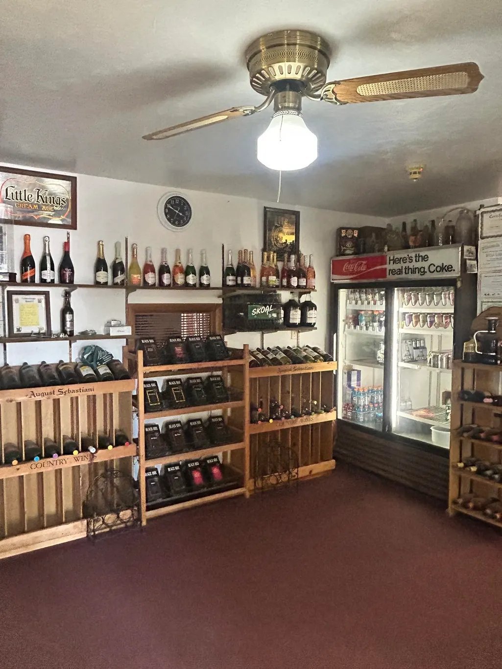 Interior of a wine shop with wooden display shelves holding wine bottles, a refrigerator case, vintage signs, and a ceiling fan