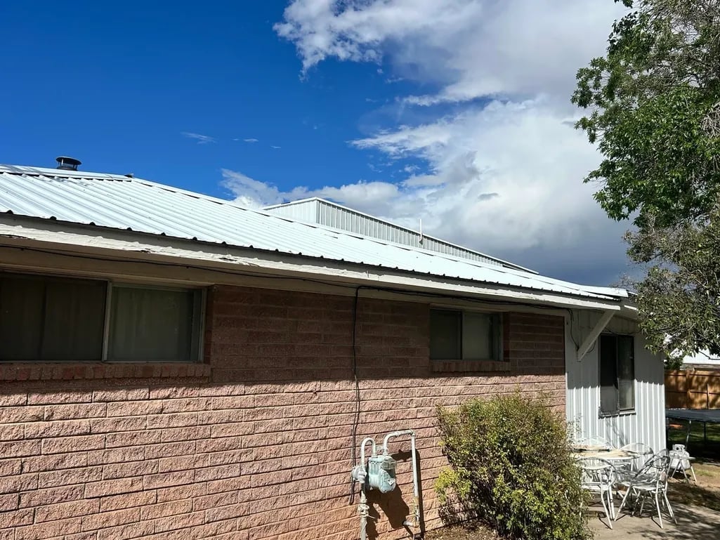 Single-story brick house with metal roof under blue sky with white clouds, featuring a porch and outdoor furniture