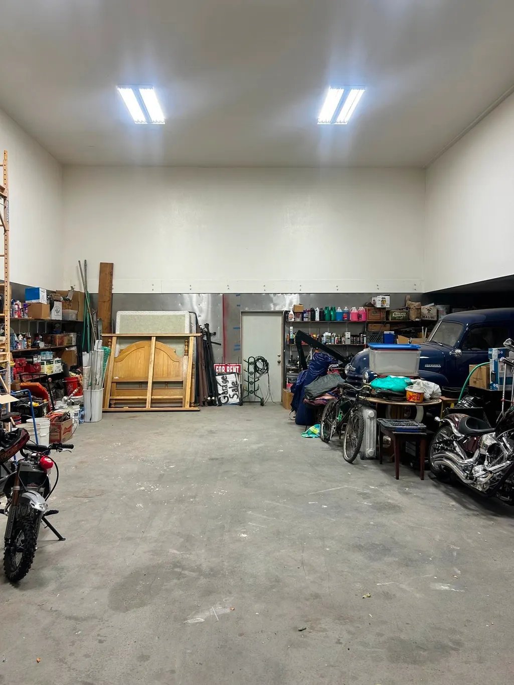 Spacious garage workshop with motorcycles, tools, storage shelves, and equipment organized along the walls under bright overhead lighting
