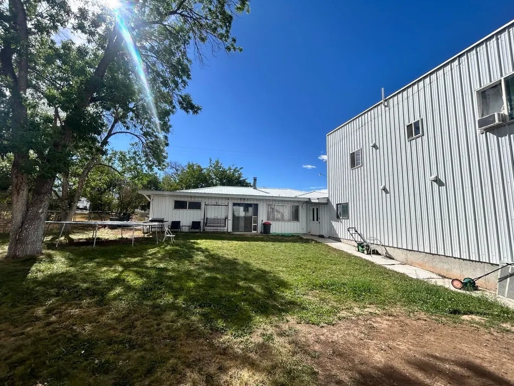 Rural property with white modular home and large metal barn building on grassy lot with mature trees under clear blue sky