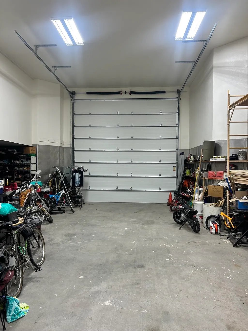Interior of a garage with a gray sectional door, concrete floor, and various bicycles and equipment stored on both sides