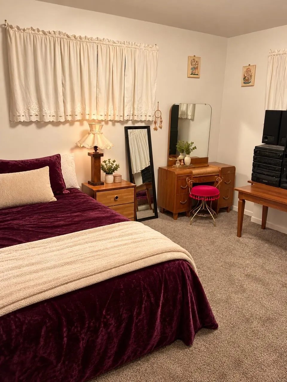 Cozy bedroom with two twin beds with burgundy velvet comforters, wooden nightstands, a vanity with mirror, and cream curtains on windows.