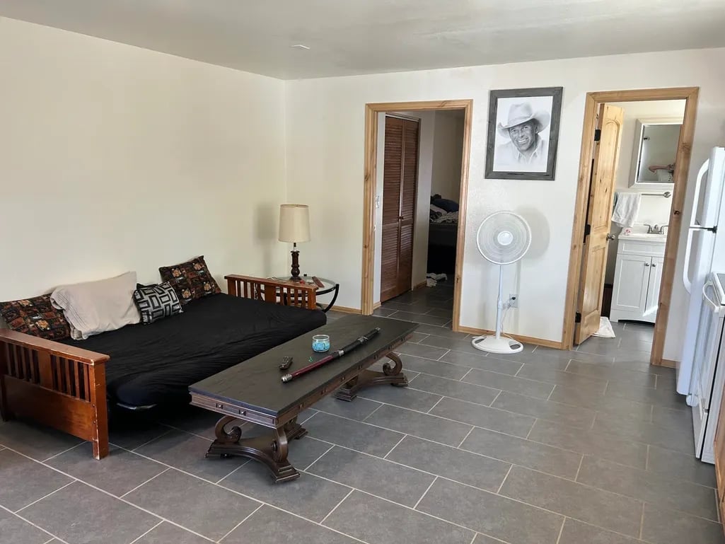 Spacious apartment interior with wooden futon, decorative pillows, coffee table, and appliances against white walls and gray tile flooring