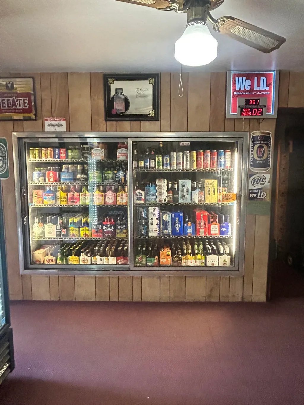 Glass-front refrigerated cooler stocked with beer and beverages in a bar setting with wood paneling and neon signs above