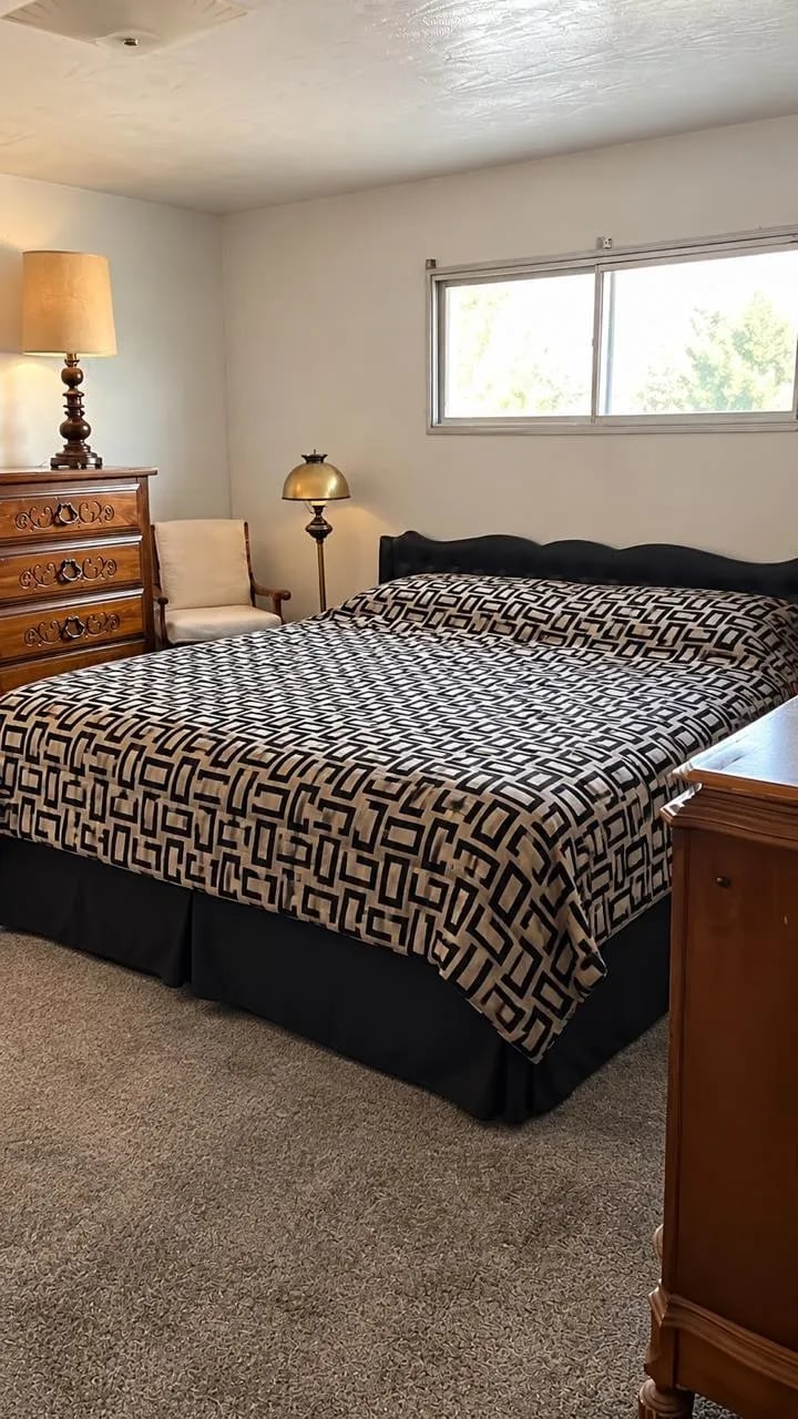 Bright bedroom with queen bed featuring geometric patterned comforter, wooden nightstands, traditional lamp, and large window overlooking greenery