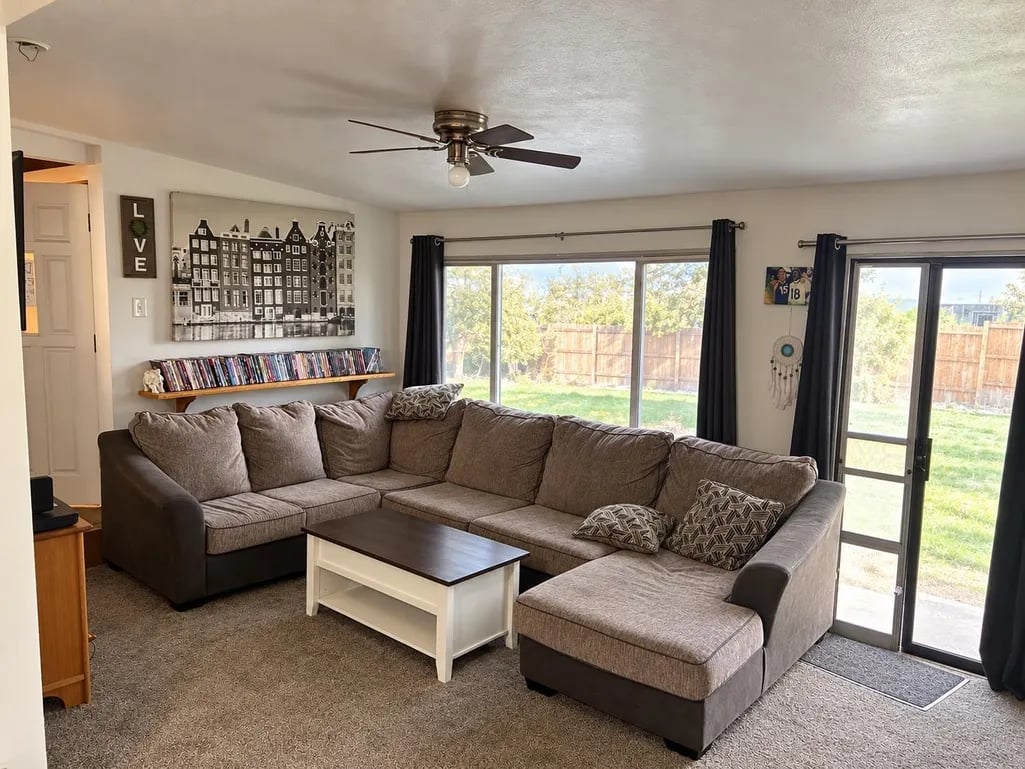 Living room with brown sectional sofa, white coffee table, ceiling fan, and glass doors opening to a backyard